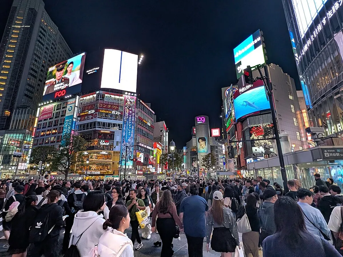 Shibuya Crossing at night, crowds streaming across the intersection beneath illuminated screens and neon signage